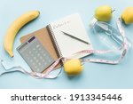 Paper notebook with word CALORIES, calculator, pen, bottle of water, measuring tape and fruits on blue background. Healthy eating concept - calculate daily nutrition intake. Top view.
