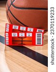 Small photo of A close-up low angle view of a pair of ticket stubs to courtside seats next to an orange basketball as it sitting a black sideline on wooden floor.
