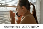 Small photo of Young scared woman looking through window on street and calling 911 police. Crime witness, spying through window, peeking on street