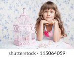 Small photo of Tender sincere dreamy romantic openhearted blond girl with long wavy hair looking at camera seating near open empty pink birdcage and lacy white table on the light blue flower background