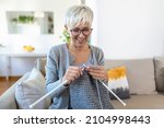 Elderly woman in glasses sit on couch at home smile holding knitting needles and yarn knits clothes for loved ones, favorite activity and pastime, retired tranquil carefree life concept
