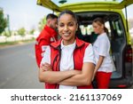 Small photo of Young female african american paramedic smiling, standing rear of the ambulance, arms crossed. Three paramedics by the ambulance