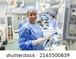 Small photo of Anesthesiologist checking monitors while sedating patient before surgical procedure in hospital operating room. Young adult female African American patient is asleep on operating table during surgery.