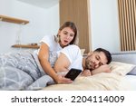 Small photo of Jealous wife spying the phone of her partner while he is sleeping in a bed at home. Shocked jealous wife spying the phone of her husband while man sleeping in bed at home