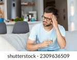 Small photo of Young man suffering from strong headache or migraine sitting with glass of water on the sofa, millennial guy feeling intoxication and pain touching aching head, morning after hangover concept