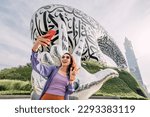 Small photo of 17 January 2023, Dubai, UAE: Tourist happy girl taking selfie photos for her travel blog, against background of the Future Museum in crescent shape and victory hand gesture