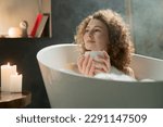 Small photo of A beautiful woman with curly hair lies in a bath with a cup of fragrant tea and looks somewhere to the side. A young woman spends her evening taking a soothing lavender oil bath and drinking delicious