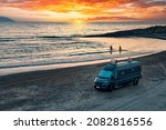 Aerial photo of campervan on abandoned beach against beutiful sunset. People bathing in the sparkling sea. Outdoor nomad lifestyle, van life holiday. Independent road trip concept.