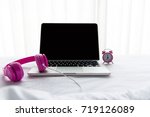 The laptop working near alarm clock and pink headphone listen music on the white bed in the sunny day morning holiday.  Music and Lifestyle Concept.