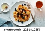 Small photo of Tiny pancakes, small thin funny crumpet, children's food. Breakfast with raspberries, blueberries, honey and nuts, white tablecloth, bright morning, top view