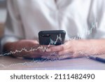 A woman trader holding in the hands a smart phone and researching stock market to proceed right investment solutions. Internet trading and wealth management concept. Hologram Forex chart.