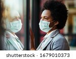 Small photo of Distraught black businesswoman wearing protective mask while standing by the window and looking through it.