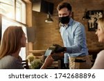 Female customer making contactless payment to a waiter who is wearing protective face mask in a cafe. 