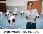 Young happy coach using stopwatch during PE class at school gym and looking at camera. Her students are exercising in the background.
