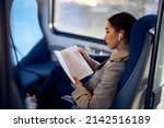 Young female passenger listening music on earbuds and reading book while traveling by train.