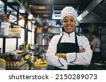 Happy African American woman standing with arms crossed while working as chef in a restaurant and looking at camera.