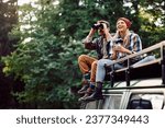 Happy woman and her boyfriend using binoculars while sitting on roof of their camper trailer and spending a day in the woods. Copy space.