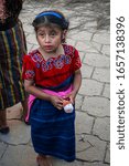 Small photo of Cajola, Quetzaltenango, Guatemala, April 27, 2019, beautiful girl in red and black typical costume, with her sad face looking at people with her toy in her hand, on the pavement road in the Mayan elec
