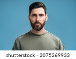 Small photo of Portrait of attentive self confident bearded man looking at camera with serious expression, unsmiling determined business man. Indoor studio shot isolated on blue background
