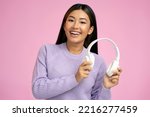 Studio portrait of relaxed cheerful asian woman holding wireless headphones isolated on pink wall. Girl enjoying listening to favorite music soundtrack 