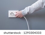 Hand plugging in an electric cord into a white plastic socket or  european wall outlet on grey plaster wall. Closeup of a woman's hand inserting an electrical plug into a wall socket. Daylight. 