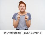 Small photo of Indoor shot of young disquieted redhead female dressed in blue striped t-shirt raising emotionally fists and biting underlip while looking discomposedly at camera, posing over white background