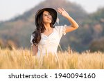 Happy young beautiful woman wearing black hat and white dress holding up her hand to block to sun shining on her face while walking in the golden barley field on a late afternoon