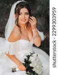 Small photo of portrait of the bride in nature. A brunette bride in a white voluminous dress is sitting, posing near a coniferous tree, holding a bouquet of white roses. Beautiful hair and makeup. Warm light