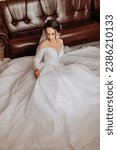 Small photo of The bride in a long, voluminous wedding dress poses sitting on the bed in her room. Long train. Portrait of the bride. Open shoulders and neckline.