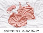 Small photo of Pink woman pajamas and sleep eye mask on white bed sheet. Top view summer pyjama for sleeping. Aesthetic lifestyle flat lay photo, singlet and shorts peach colored, comfort ans stylish wear or clothes