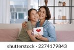Small photo of Happy time Mother day grown up child girl looking at camera cuddle hug give gift box heart card to mature mum. Love kiss care mom asia middle age adult people smile enjoy relax sitting at home sofa.