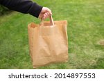 Small photo of Close up of man hand with eco paper bag for takeaway food on nature green background. Delivery in any weather around the clock to the client.