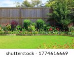 Colourful Pretty Flower And Shrub Border Surrounded By A Fence And A Green Lawn In A Home Back Garden.