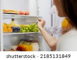 Woman hand taking, grabbing or picks up green bunch of grapes out of open refrigerator shelf or fridge drawer full of fruits, vegetables, banana, peaches, yogurt. Healthy food diet, lifestyle