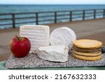 Small photo of Four famous cheeses of Normandy, squared pont l'eveque, round camembert cow cheese, yellow livarot, heartshaped neufchatel and view on promenade and alebaster cliffs in Etretat, Normandy, France