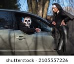 Small photo of A man in the guise of a dead man in a car and a nun- witch near the hood of a car on Halloween.