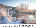 Winter landscape of sunrise in the woodland covered snow. Mist over river witout ice. Dry grass covered snow. Tops trees lighted rays sun in the morning. 