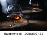 Small photo of Glasses of cognac of amber color stand on barrels of cognac in wine cellar illuminated by soft light. Brandy in glasses on wooden barrel of amber-colored alcohol.