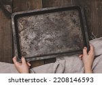 Two female hands hold a rectangular empty baking sheet on a brown wooden table, top view