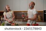 Small photo of Happy Caucasian senior old couple having fun cooking at home funny man fooling around juggling tomatoes smiling woman cook fresh vegetable salad in kitchen playful family juggle natural food products