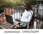 Small photo of A portrait of a young drop-dead gorgeous black woman freelancer with long braided hair and white shirt, having a morning coffee and breakfast on the balcony of her house and lazily using the laptop