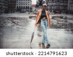 Small photo of View of a young cute black female in the demi-season overcoat, with short hair, painted white, standing on a wet city square after the rain and leaning against her closed umbrella, Lisbon, Portugal