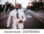 Small photo of A stylish black man with a bald head, long beard, and hints of white hair, dressed in a pink wrap jacket, holds a cup of coffee with contentment semblance in the middle of a bicycle lane in Lisbon