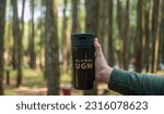 Small photo of "Alumni UGM" engraved on a Tumblr glass, held by a hand against a pine forest backdrop. Pride and nostalgia resonate in this captivating image.