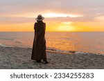 Lonely pensive woman walking at sunset by sea, enjoying nature. Rear view of unrecognizable stylish senior lady standing alone on seashore against yellow sky, copy space.