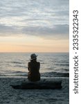 Small photo of Back view of pensive lonely woman in coat and hat sitting on log on seashore looking away at sea in evening.