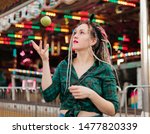 Small photo of Inordinate young woman with dreadlocks hairstyle and fashionable summer clothes throws apple in amusement park.