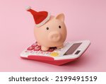 Christmas piggy bank in santa hat and calculator on pink background