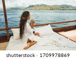 Woman enjoying morning coffee on boat in private cruise tour. Bed on board. Travel and freedom lifestyle. Mountain view of Komodo national park on horizon.