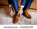 A man ties up his shoelaces on his brown shoes in the room. Blue suit and patent leather shoes. 
